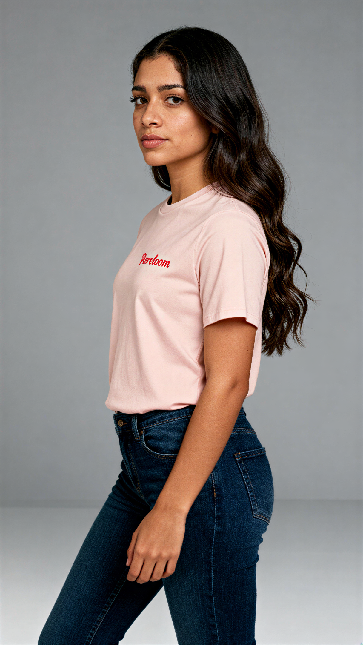 במבט צדדי, אישה לובשת חולצת כותנה אורגנית בצבע ורוד עם לוגו אדום "Pureloom" בצד שמאל של החזה מבית פיורלום.